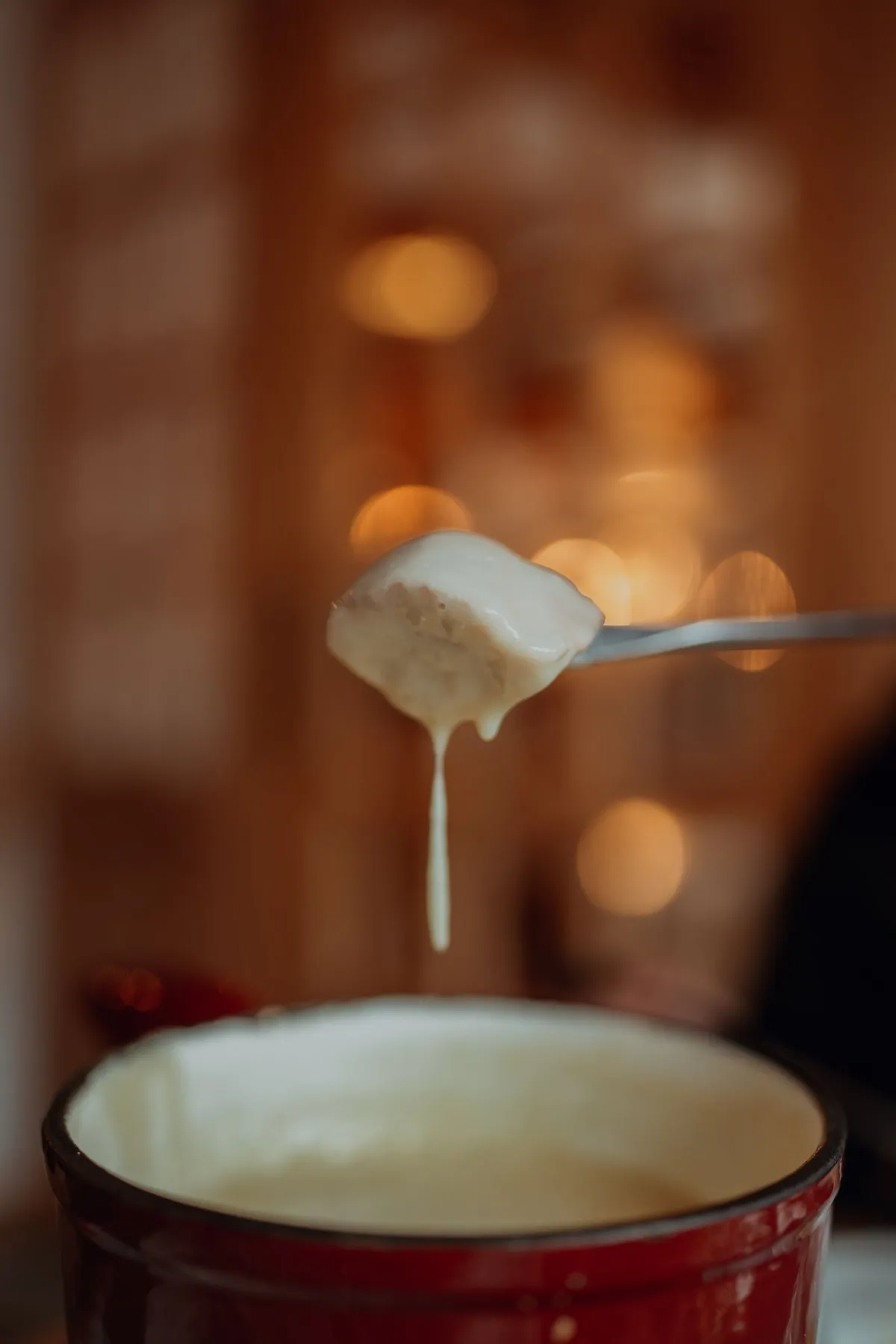 A fondue fork lifting a bread cube with a long drip of melted cheese over a red pot